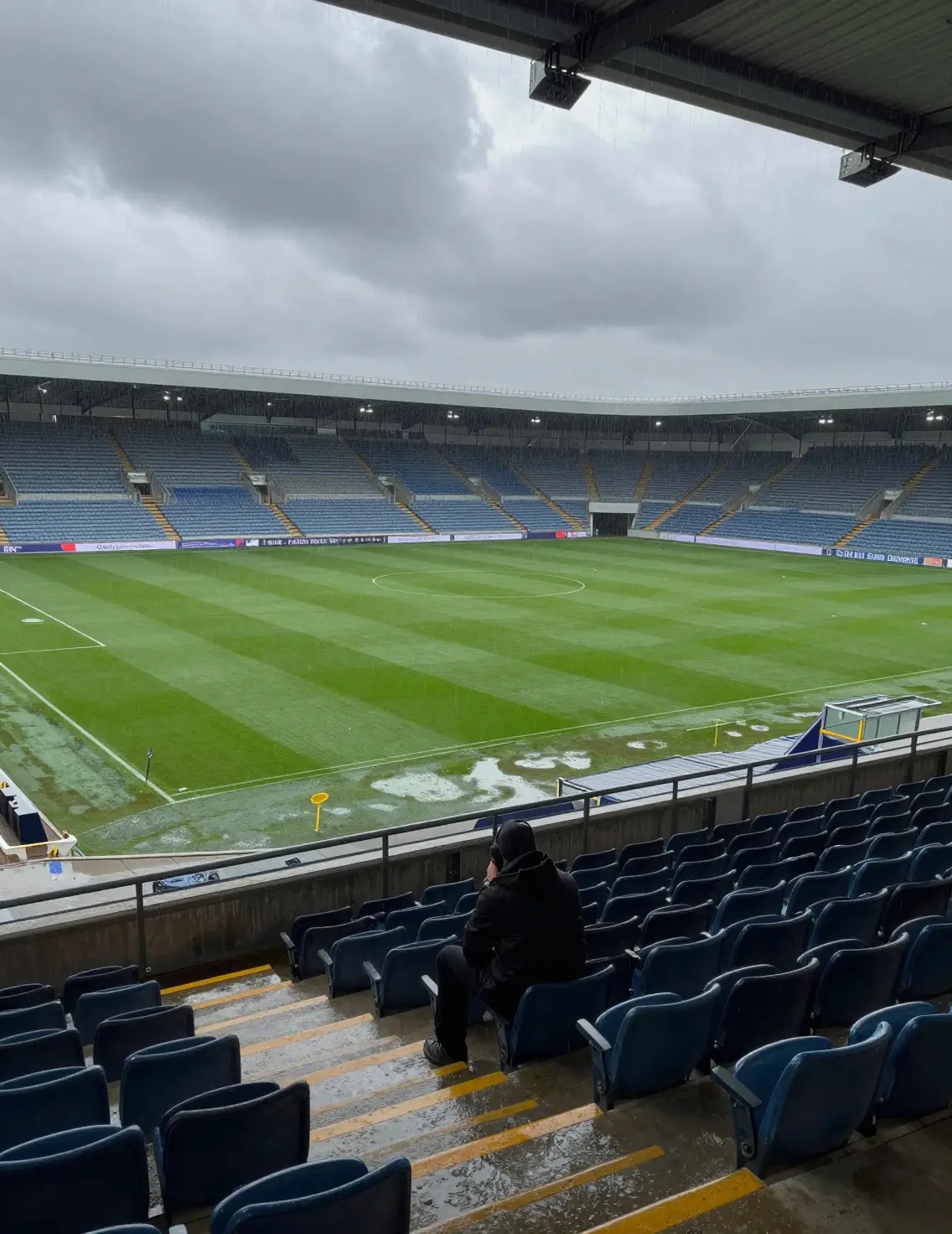 Un unico spettatore siede sopra un campo da calcio vuoto e fradicio di pioggia